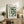 May include: Framed Bauhaus modernist poster with green geometric semicircles and "MODERN ART" text, displayed on rustic wooden sideboard beside fern plant, ceramic vase, and books in neutral-toned living room with beige couch and warm lighting