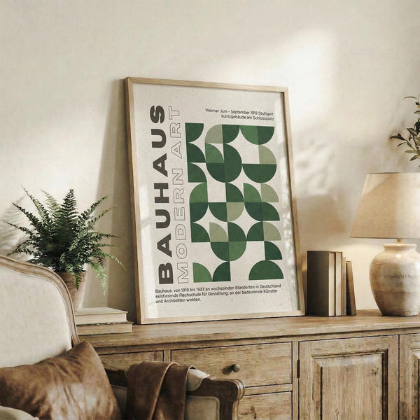 May include: Framed Bauhaus modernist poster with green geometric semicircles and "MODERN ART" text, displayed on rustic wooden sideboard beside fern plant, ceramic vase, and books in neutral-toned living room with beige couch and warm lighting