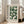 May include: Framed Bauhaus modernist poster with geometric green semicircles and quarter-circles arranged in a grid pattern on cream background, displayed in light wood frame leaning against white wall next to beige upholstered chair in bright living room with tall windows and natural lighting.