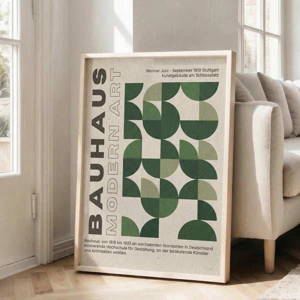 May include: Framed Bauhaus modernist poster with geometric green semicircles and quarter-circles arranged in a grid pattern on cream background, displayed in light wood frame leaning against white wall next to beige upholstered chair in bright living room with tall windows and natural lighting.