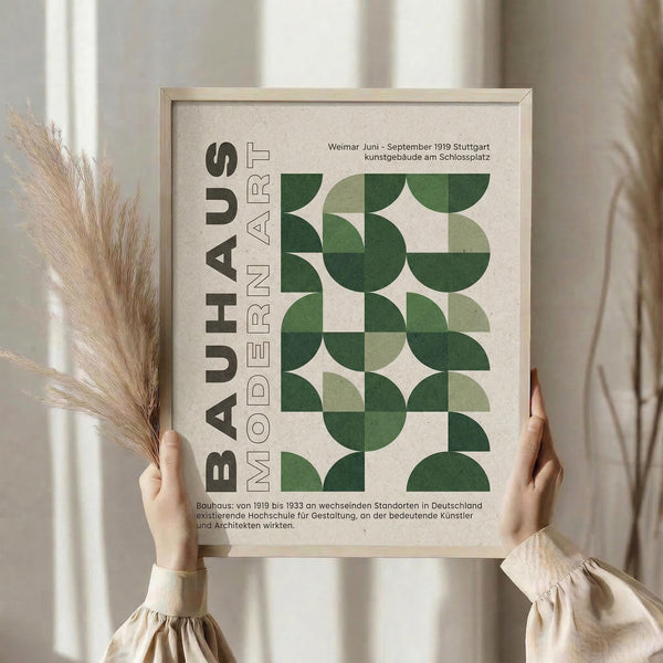May include: Person holding framed Bauhaus-inspired poster with geometric green circles and semicircles in various shades, arranged in grid pattern on cream background. German text includes "BAUHAUS MODERN ART" and historical information. Light wood frame, dried wheat stalks visible, neutral interior setting with soft natural lighting.