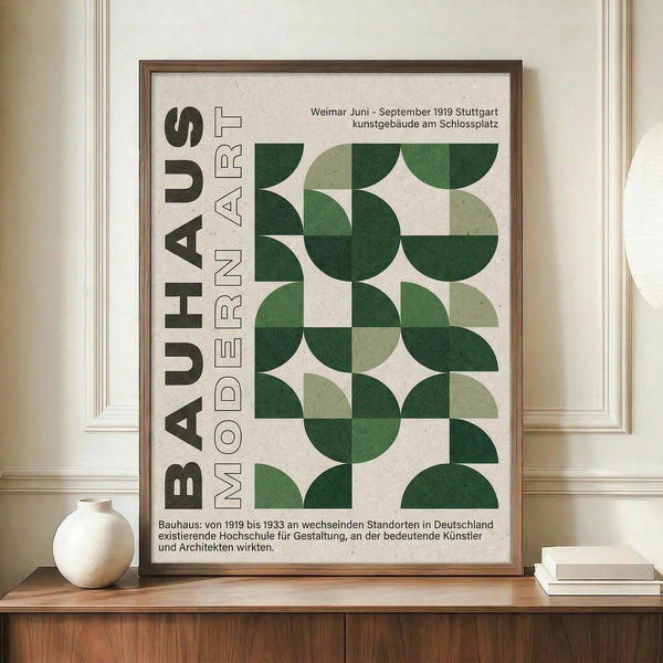 May include: Framed Bauhaus-style poster with green geometric semicircles arranged in a grid pattern on cream background, displayed on wooden sideboard next to white ceramic vase and stacked books in room with white paneled walls and warm lighting.