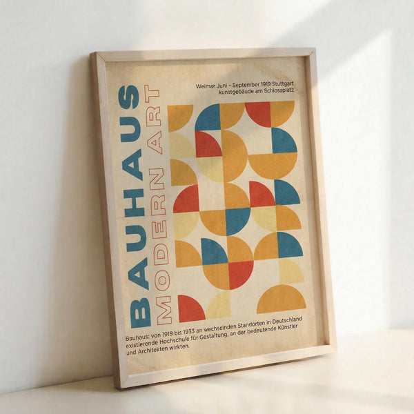 May include: A framed poster with geometric design featuring colorful semicircles and quarter circles in red, orange, blue and yellow arranged in a grid pattern. Text reads "BAUHAUS MODERN ART" vertically on left side, with German text about Weimar 1919 Stuttgart exhibition. Light wood frame leaning against white wall on white surface.