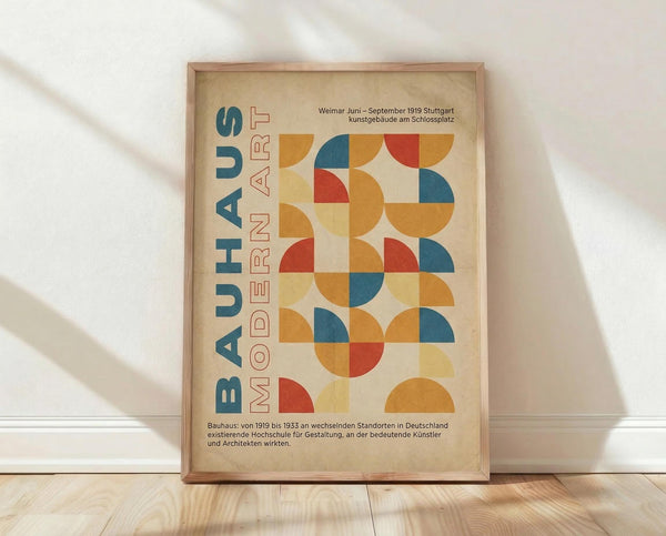 May include: Framed Bauhaus-inspired poster with geometric pattern of colorful semicircles and quarter-circles in orange, blue, red, and yellow on cream background. German text reads "BAUHAUS MODERN ART" vertically. Wooden frame rests against white wall on light wood flooring with soft shadows from window light.