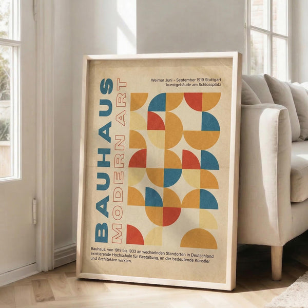 May include: A framed Bauhaus-style poster with geometric semicircles in red, orange, blue and yellow arranged in a grid pattern. The cream-colored print features "BAUHAUS" and "MODERN ART" text along the left side, with German exhibition details at top and bottom. The white wooden frame leans against a white wall beside a beige upholstered chair in a bright room with large windows.