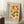 May include: Framed Bauhaus modern art poster leaning against white wall corner next to beige armchair and wooden side table with coffee cup. Poster features colorful geometric circles in yellow, blue, red, and orange with German text. Natural lighting creates soft shadows on white fluffy rug below.