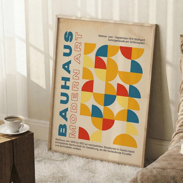 May include: Framed Bauhaus modern art poster leaning against white wall corner next to beige armchair and wooden side table with coffee cup. Poster features colorful geometric circles in yellow, blue, red, and orange with German text. Natural lighting creates soft shadows on white fluffy rug below.