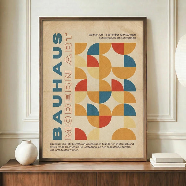 May include: Framed Bauhaus-style poster with geometric circles and semicircles in orange, red, blue, and yellow arranged in a grid pattern. German text reads "BAUHAUS MODERN ART" vertically on left side. Cream-colored background with vintage texture. Dark wood frame displayed on wooden sideboard next to white ceramic vase and stacked books in bright room with white walls and molding.