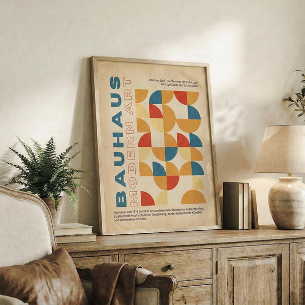 May include: Framed Bauhaus-inspired poster with geometric circles and semicircles in orange, blue, red, and yellow tones, displayed on rustic wooden sideboard alongside fern plant, pottery vase, stacked books, and leather armchair in neutral-toned living room setting