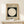 May include: Framed Bauhaus poster with white circle on black square background, displayed on white dresser next to small wrapped gift with sprig. Light wood frame, German text below geometric design, soft natural lighting on cream wall.