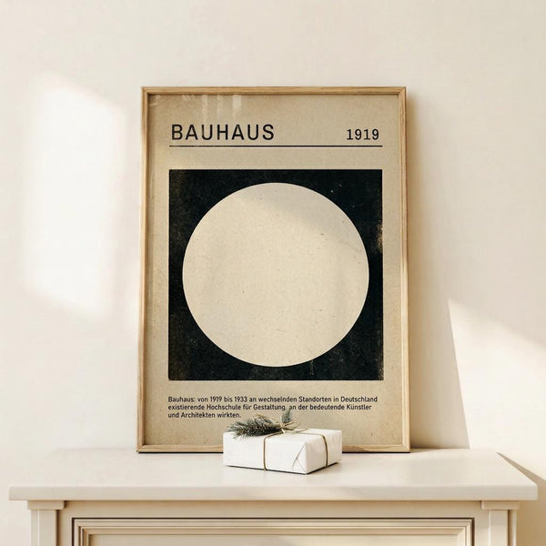 May include: Framed Bauhaus poster with white circle on black square background, displayed on white dresser next to small wrapped gift with sprig. Light wood frame, German text below geometric design, soft natural lighting on cream wall.