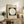 May include: Framed Bauhaus poster with white circle on dark green square background, dated 1919, displayed on rustic wooden sideboard alongside fern plant, ceramic vases, and books in neutral-toned living room with beige armchair and white walls