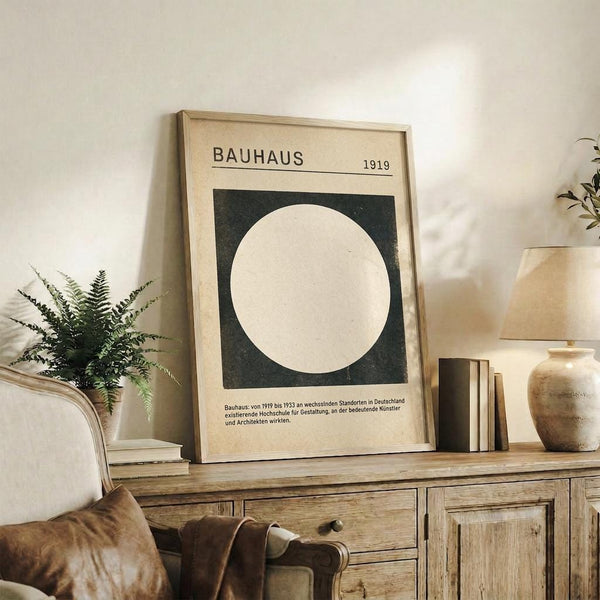 May include: Framed Bauhaus poster with white circle on dark green square background, dated 1919, displayed on rustic wooden sideboard alongside fern plant, ceramic vases, and books in neutral-toned living room with beige armchair and white walls