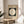 May include: Framed Bauhaus poster from 1919 featuring a large white circle on dark green background with cream matting and thin wood frame, displayed on round wooden stool beside beige upholstered chair and plant, with natural sunlight streaming across white walls creating geometric shadows