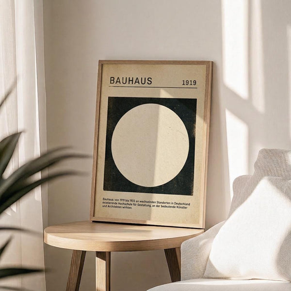 May include: Framed Bauhaus poster from 1919 featuring a large white circle on dark green background with cream matting and thin wood frame, displayed on round wooden stool beside beige upholstered chair and plant, with natural sunlight streaming across white walls creating geometric shadows