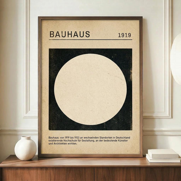 May include: Framed Bauhaus-style poster with cream background featuring large white circle on black square, dated 1919, with German text below. Displayed on wooden sideboard next to white ceramic vase and stacked books against cream wainscoted wall.