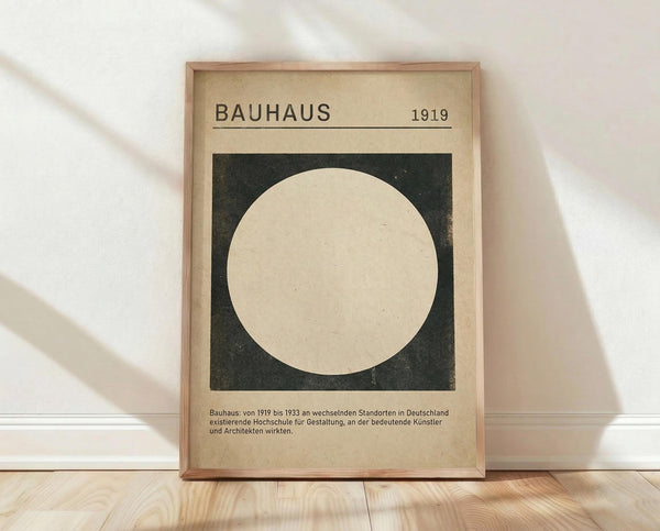 May include: Framed Bauhaus poster with beige background and dark green square containing large cream circle, displayed on light wood floor against white wall with natural lighting creating soft shadows across the scene