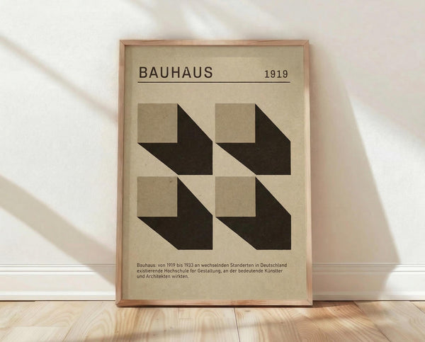 May include: Framed Bauhaus-style poster with geometric design featuring four black arrow-like shapes pointing inward on cream background. Text reads "BAUHAUS 1919" at top with German description below. Light wood frame leaning against white wall on natural wood flooring with soft natural lighting casting shadows.