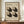 May include: Framed Bauhaus-style poster with geometric brown and beige squares creating dimensional pattern, displayed on wooden sideboard next to white ceramic vase and stacked books against cream paneled wall in modern interior setting.