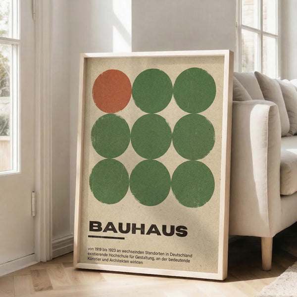 May include: A framed Bauhaus-style poster featuring nine circles in a 3x3 grid - one rust orange circle in the top left, eight green circles filling the remaining positions. Bold black "BAUHAUS" text below with German description text. Cream-colored background with visible texture. Light wood frame leaning against white wall next to beige upholstered sofa in bright, airy living room with large windows.