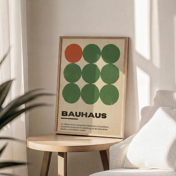 May include: Framed Bauhaus poster with nine circles arranged in a 3x3 grid - one orange circle in top left, eight green circles filling remaining positions, German text below on cream background. Wooden frame sits on round wooden stool beside beige armchair with throw pillow, plant leaves visible, soft natural lighting creates shadows on white wall.