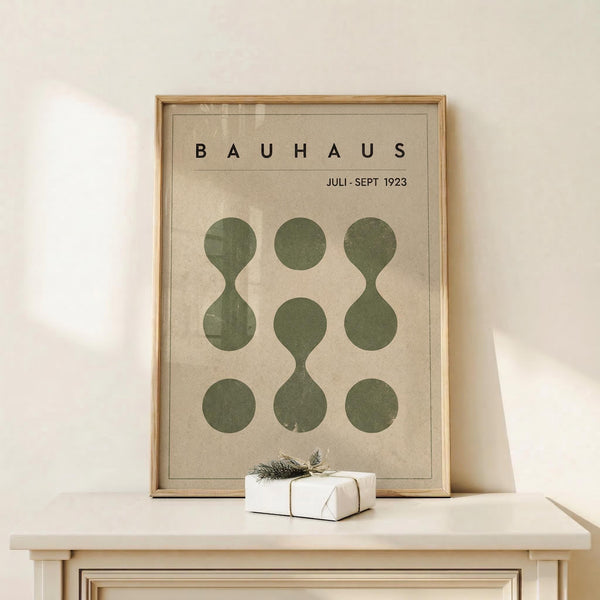 May include: Framed Bauhaus poster with beige background displaying nine olive green circles arranged in 3x3 grid, some connected to form hourglass shapes. Black text reads "BAUHAUS" and "JULI-SEPT 1923" at top. Natural wood frame sits on white wooden console table beside small wrapped gift with evergreen sprig. White wall background with soft natural lighting creates minimalist scandinavian-style room setting.