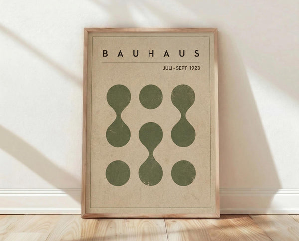 May include: Framed Bauhaus-style poster with beige background featuring nine olive green circles arranged in a 3x3 grid, some connected with flowing curved lines. Black text reads "BAUHAUS" and "JULI-SEPT 1923" at top. Natural wood frame sits on light wooden floor against white wall with soft window light casting shadows.