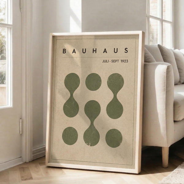 May include: Framed vintage-style Bauhaus poster with beige background and nine olive green circles arranged in a 3x3 grid, some circles connected to form hourglass or figure-eight shapes. Light wood frame leaning against white wall next to beige sofa in bright, minimalist living room with large windows and wooden floors.