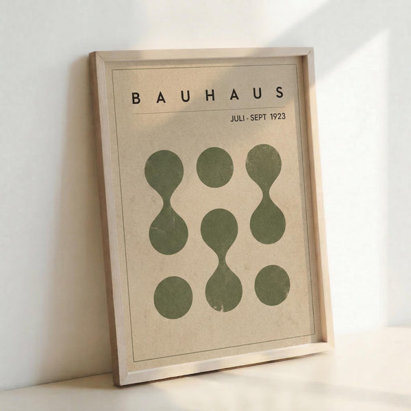 May include: A framed Bauhaus-style poster print leaning against a white wall. The artwork features nine olive green circles arranged in a 3x3 grid on a cream-colored background, with some circles connected to form hourglass or figure-eight shapes. Black text reads "BAUHAUS" and "JULI-SEPT 1923" at the top. The minimalist design is displayed in a light wood frame, casting soft shadows on the wall behind it.