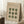 May include: Framed Bauhaus-style poster with beige background and dark green geometric circles arranged in a 3x3 grid, some connected to form hourglass shapes. Text reads "BAUHAUS" and "JULI-SEPT 1923" at top. Poster leans against white wall corner on white fluffy rug, with wooden stool holding white coffee cup and books nearby, and beige throw blanket visible.