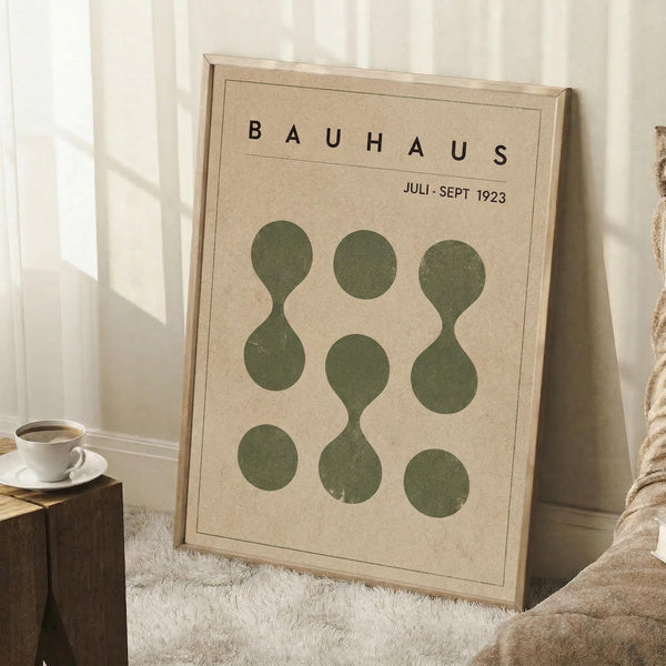 May include: Framed Bauhaus-style poster with beige background and dark green geometric circles arranged in a 3x3 grid, some connected to form hourglass shapes. Text reads "BAUHAUS" and "JULI-SEPT 1923" at top. Poster leans against white wall corner on white fluffy rug, with wooden stool holding white coffee cup and books nearby, and beige throw blanket visible.
