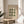 May include: Framed Bauhaus exhibition poster with green circular and oval shapes arranged in geometric pattern on cream background, displayed on light wood round side table next to beige upholstered chair, with plant leaves visible and natural window light creating soft shadows on neutral wall.