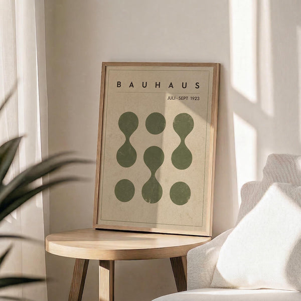 May include: Framed Bauhaus exhibition poster with green circular and oval shapes arranged in geometric pattern on cream background, displayed on light wood round side table next to beige upholstered chair, with plant leaves visible and natural window light creating soft shadows on neutral wall.
