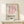 May include: Pink and white curved geometric design resembling letter D on cream background with black "BAUHAUS" text and German description below, displayed in thin gold frame on white wooden console table next to small wrapped gift with twine and greenery sprig against white wall