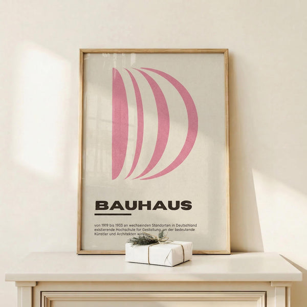 May include: Pink and white curved geometric design resembling letter D on cream background with black "BAUHAUS" text and German description below, displayed in thin gold frame on white wooden console table next to small wrapped gift with twine and greenery sprig against white wall