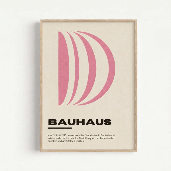 A framed poster with pink curved shapes forming a letter D on cream background, displayed against a white wall. Below the geometric design is bold black text reading "BAUHAUS" with an underline, followed by German text describing the design school that operated from 1919 to 1933 at various locations in Germany. The artwork is housed in a light wood frame and showcases minimalist Bauhaus design principles with clean typography and simple geometric forms.