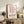 May include: Framed pink geometric poster with curved lines and Bauhaus text displayed on rustic wooden sideboard beside cream armchair, fern plant, ceramic vases, and stacked books in warm, naturally lit living room setting