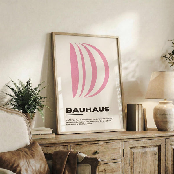 May include: Framed pink geometric poster with curved lines and Bauhaus text displayed on rustic wooden sideboard beside cream armchair, fern plant, ceramic vases, and stacked books in warm, naturally lit living room setting