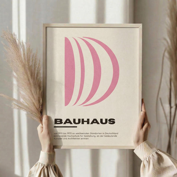 May include: Person holding framed pink Bauhaus poster featuring curved geometric shapes in soft pink tones on cream background. Light wood frame, minimalist design with German text below. Dried pampas grass visible to left, neutral beige clothing and curtains create warm, modern interior styling.