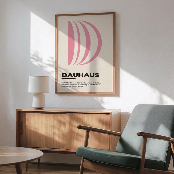 May include: Pink geometric curved shapes forming letter D on cream background in thin wooden frame, hung above mid-century modern wooden credenza with white ceramic table lamp, next to sage green upholstered armchair with wooden arms in bright sunlit room with white walls and hardwood floors.
