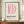 May include: A framed poster displaying curved pink shapes resembling a stylized "D" above bold "BAUHAUS" text and German description text, shown in a thin brown wooden frame on a light wooden surface alongside a cream ceramic vase and white books, against cream wainscoted walls.