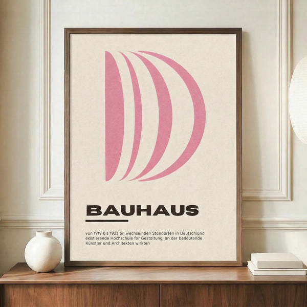 May include: A framed poster displaying curved pink shapes resembling a stylized "D" above bold "BAUHAUS" text and German description text, shown in a thin brown wooden frame on a light wooden surface alongside a cream ceramic vase and white books, against cream wainscoted walls.