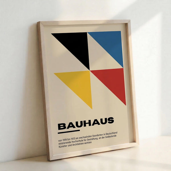 Framed poster featuring geometric design with four triangular shapes in black, blue, yellow, and red against cream background. Bold "BAUHAUS" text below with German description text. Light wood frame leaning against white wall, casting subtle shadow.