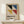 May include: Framed Bauhaus poster with geometric design featuring four colored triangles - black, blue, yellow, and red - arranged around white diagonal stripes on beige background. German text below describes the design school's history. Natural wood frame sits on light wooden shelf next to small white wrapped gift with twine and pine sprig. White wall backdrop with soft natural lighting creates minimalist display setting.