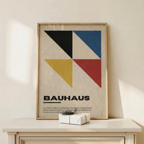 May include: Framed Bauhaus poster with geometric design featuring four colored triangles - black, blue, yellow, and red - arranged around white diagonal stripes on beige background. German text below describes the design school's history. Natural wood frame sits on light wooden shelf next to small white wrapped gift with twine and pine sprig. White wall backdrop with soft natural lighting creates minimalist display setting.