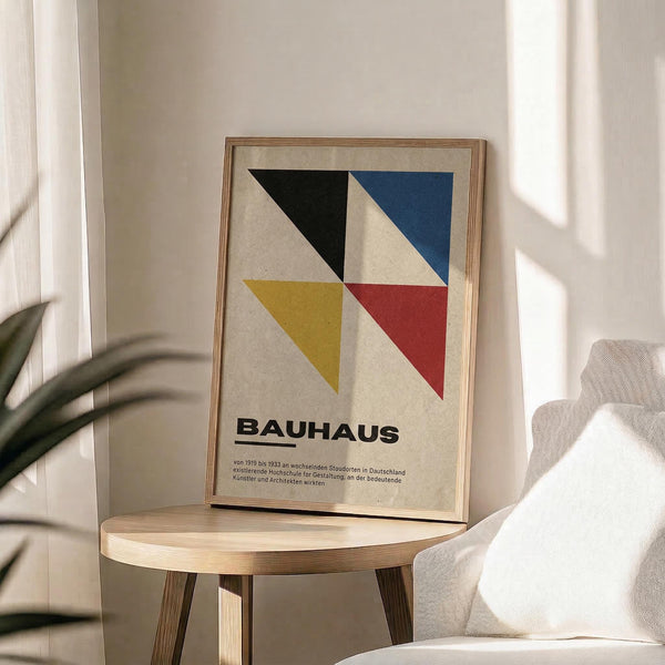 May include: Framed Bauhaus poster with geometric design featuring black, blue, yellow, and red triangular shapes on cream background, displayed on light wood round side table next to beige upholstered chair. Natural sunlight streams across neutral-toned room with plant leaves visible in corner, creating soft shadows on white wall.