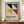 May include: Framed Bauhaus poster with geometric design featuring black, blue, yellow, and red triangular shapes on beige background, displayed on wooden sideboard next to white ceramic vase and books in elegant room with white paneled walls
