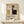 May include: Framed Bauhaus poster with geometric abstract design featuring colored rectangles in red, blue, yellow, and black on beige background, displayed on light wood surface next to small wrapped gift with twine and evergreen sprig against white wall