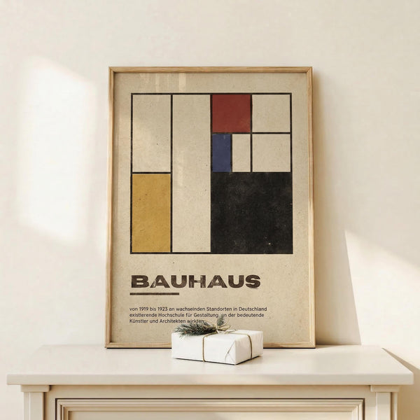 May include: Framed Bauhaus poster with geometric abstract design featuring colored rectangles in red, blue, yellow, and black on beige background, displayed on light wood surface next to small wrapped gift with twine and evergreen sprig against white wall