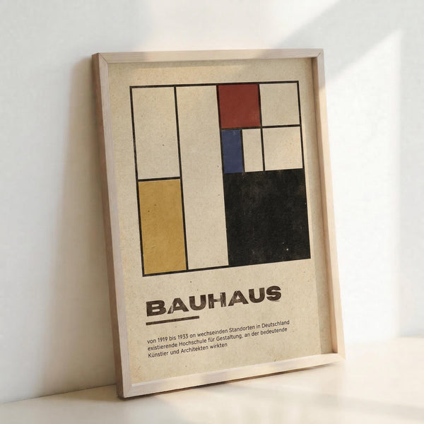 Framed poster featuring a geometric abstract composition with colored rectangles in yellow, red, blue, and black against cream background, displayed in light wooden frame leaning against white wall. German text below reads "BAUHAUS" with smaller descriptive text about the design school that operated from 1919-1933 in various German locations.